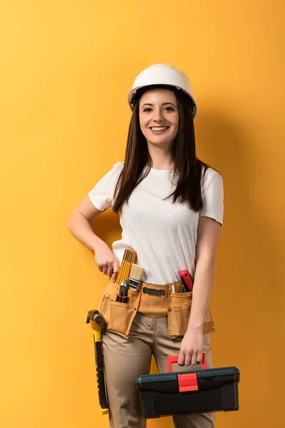 Sonriente manitas en casco sosteniendo caja de herramientas sobre fondo amarillo - foto de stock