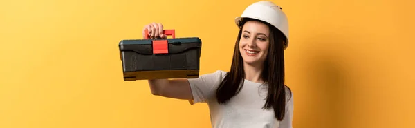 Panoramic shot of smiling handywoman in helmet holding toolbox on yellow background — Stock Photo