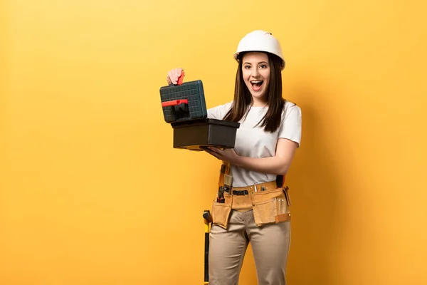 Shocked handywoman in helmet holding toolbox on yellow background — Stock Photo