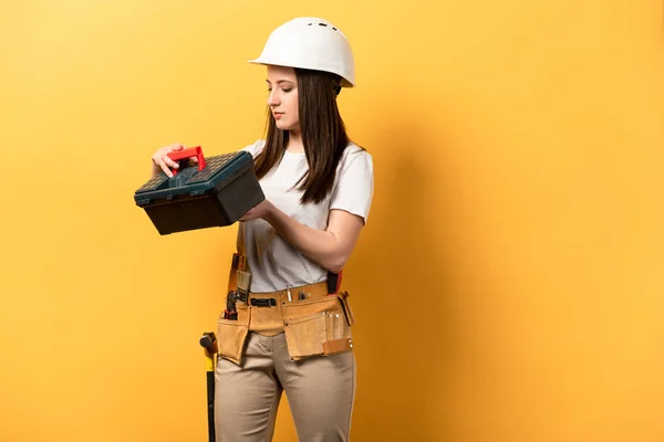 Attractive handywoman in helmet holding toolbox on yellow background — Stock Photo