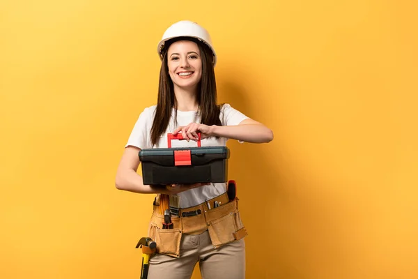 Smiling handywoman in helmet holding toolbox on yellow background — Stock Photo