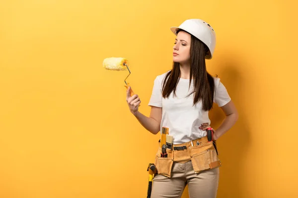Серьезная женщина в шлеме держа ролик краски на желтом фоне — стоковое фото