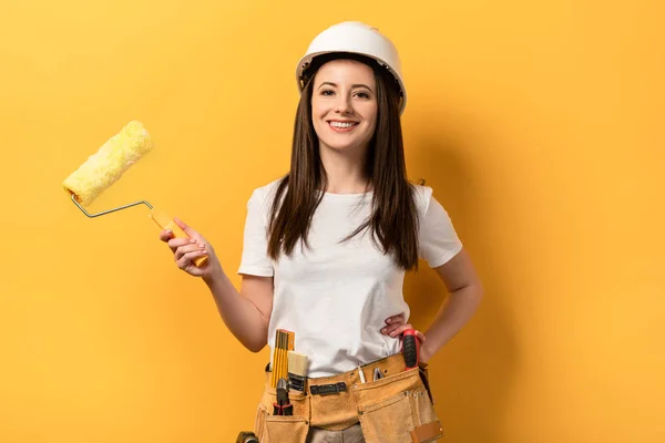 Smiling handywoman with hand on hip holding paint roller on yellow background — Stock Photo