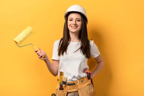 Sorrindo handywoman com a mão no quadril segurando rolo de pintura no fundo amarelo — Fotografia de Stock