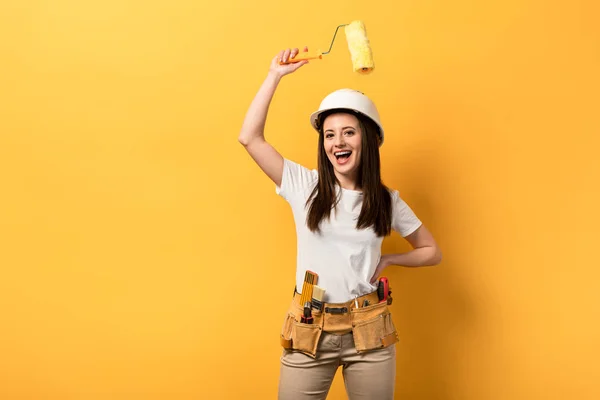 Sorrindo handywoman com a mão no quadril segurando rolo de pintura no fundo amarelo — Fotografia de Stock