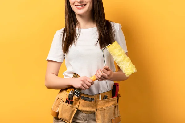 Vista recortada de manitas sonrientes sosteniendo rodillo de pintura sobre fondo amarillo - foto de stock