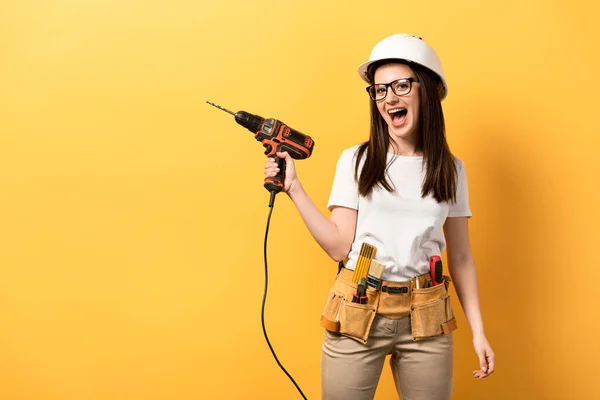 Positive handywoman screaming and holding drill on yellow background — Stock Photo