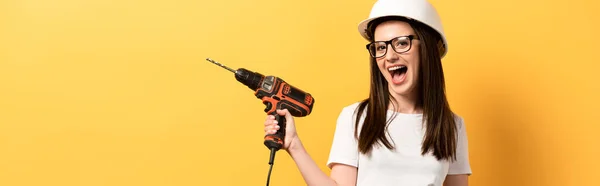 Panoramic shot of positive handywoman screaming and holding drill on yellow background — Stock Photo