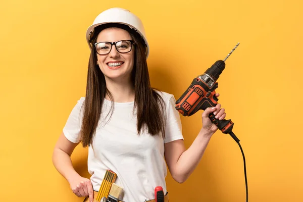 Lächelnde Handarbeiterin im Helm mit Bohrmaschine auf gelbem Hintergrund — Stockfoto