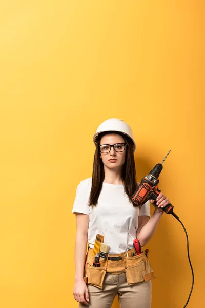 Sérieuse bricoleuse tenant foret et regardant la caméra sur fond jaune — Photo de stock