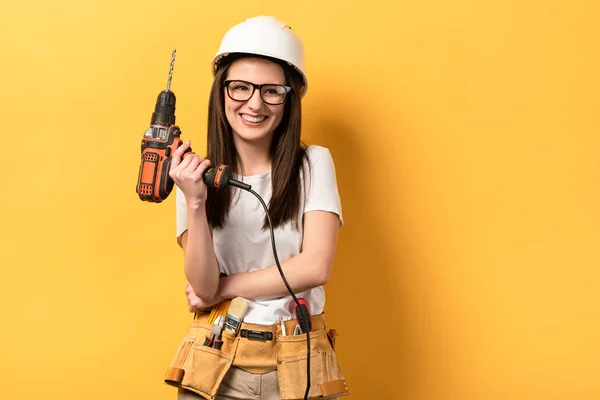 Lächelnde Handarbeiterin im Helm mit Bohrmaschine auf gelbem Hintergrund — Stockfoto