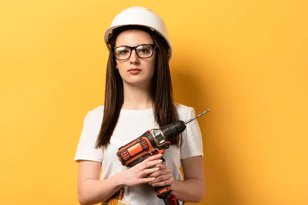 Sérieuse bricoleuse tenant foret et regardant la caméra sur fond jaune — Photo de stock