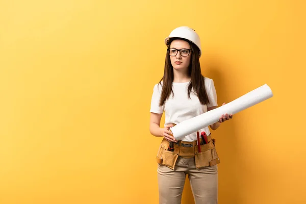 Serious handywoman holding blueprint and looking away on yellow background — Stock Photo
