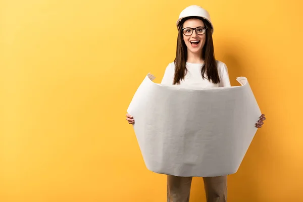 Shocked handywoman holding blueprint on yellow background with copy space — Stock Photo