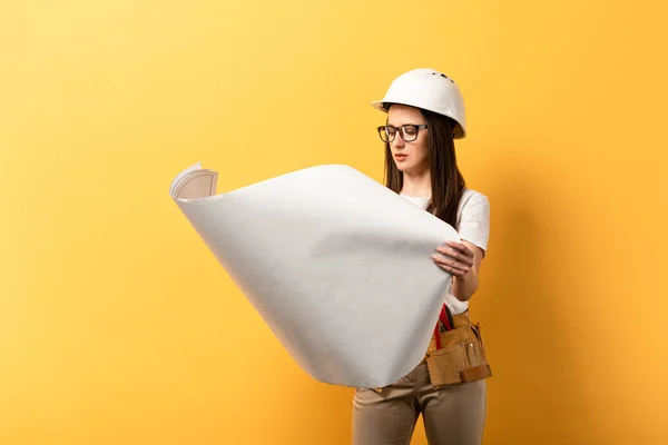 Attractive handywoman in helmet looking at plan on yellow background — Stock Photo