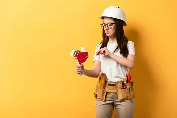 Bricoleuse dans le casque tenant distributeur de ruban sur fond jaune — Photo de stock