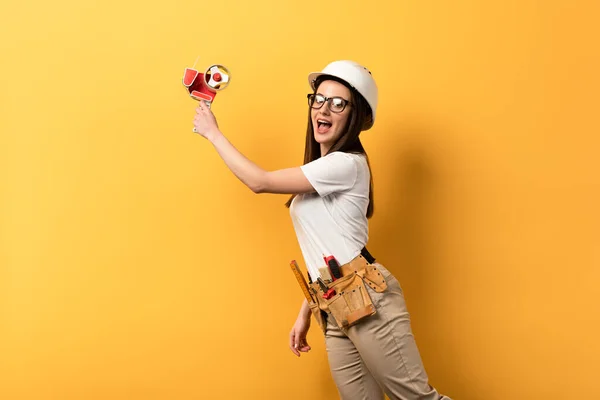 Chocado handywoman segurando dispensador de fita no fundo amarelo com espaço de cópia — Fotografia de Stock