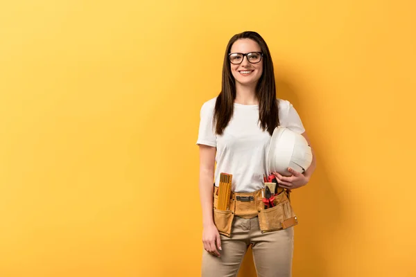 Smiling handywoman holding helmet and looking at camera on yellow background — Stock Photo