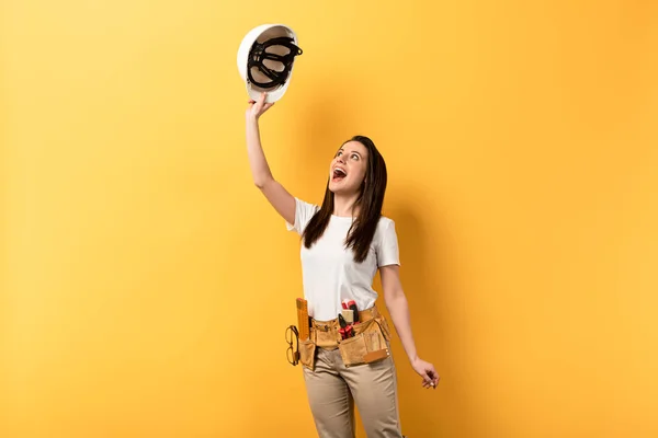 Surpresa handywoman segurando e olhando para o capacete no fundo amarelo — Fotografia de Stock