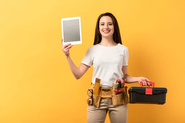 Lächelnde Handarbeiterin mit digitalem Tablet und Werkzeugkiste auf gelbem Hintergrund — Stockfoto