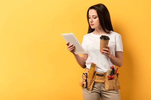 Handywoman usando tablet digital e segurando copo de papel no fundo amarelo — Fotografia de Stock