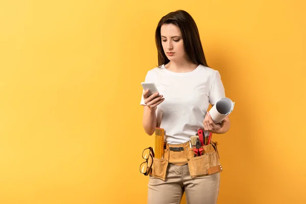 Serious handywoman using smartphone and holding blueprint on yellow background — Stock Photo