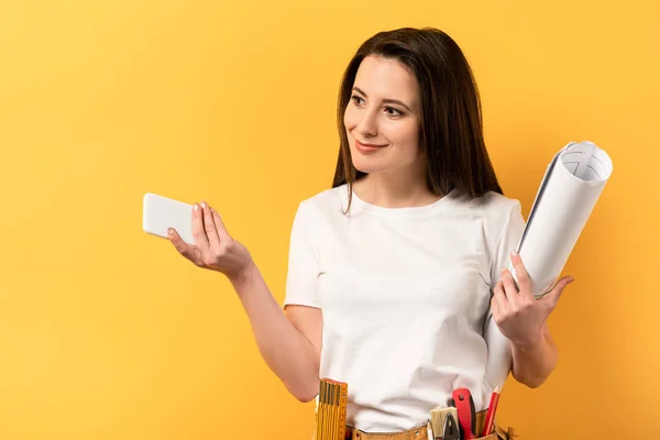 Sorrindo handywoman segurando smartphone e blueprint no fundo amarelo — Fotografia de Stock