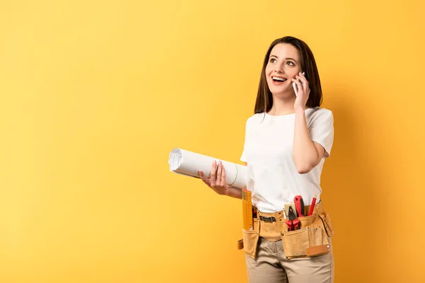 Smiling handywoman holding smartphone and blueprint on yellow background — Stock Photo