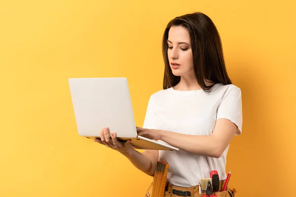 Handywoman atraente usando e segurando laptop no fundo amarelo — Fotografia de Stock