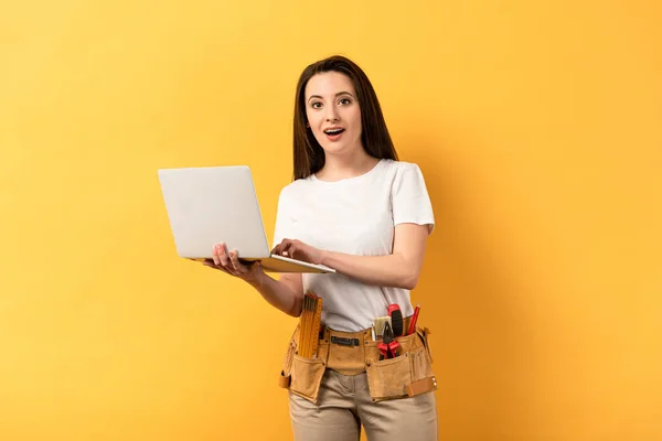 Shocked repairwoman holding laptop and looking at camera on yellow background — Stock Photo