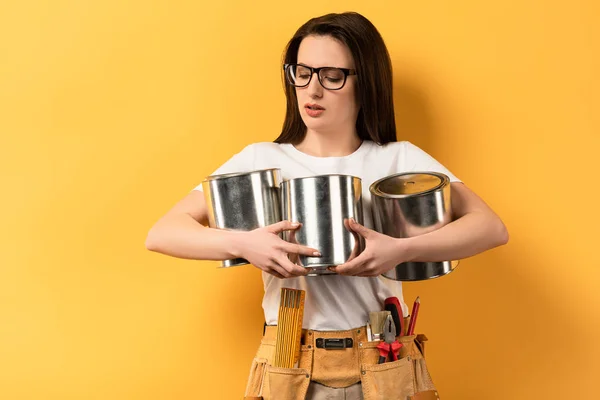 Attractive repairwoman holding and looking at paint cans on yellow background — Stock Photo