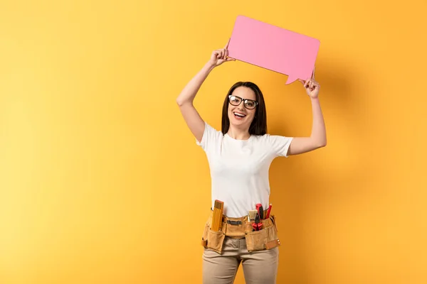 Smiling repairwoman holding speech bubble and looking at camera on yellow background — Stock Photo