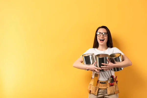 Schockierte Reparaturfrau hält Farbdosen in der Hand und schaut auf gelbem Hintergrund weg — Stockfoto