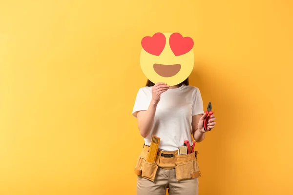 KYIV, UKRAINE - DECEMBER 16, 2019: repairwoman obscuring face with smiley with heart eyes and holding pliers on yellow background — Stock Photo