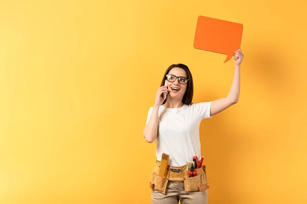 Smiling repairwoman holding speech bubble and talking on smartphone on yellow background — Stock Photo