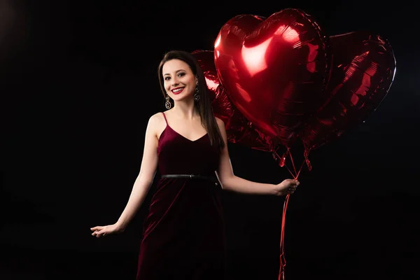 Mulher sorridente em vestido segurando balões em forma de coração em 14 de fevereiro isolado em preto — Fotografia de Stock