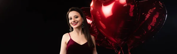 Panoramic shot of smiling woman in dress holding heart-shaped balloons in 14 february isolated on black — Stock Photo
