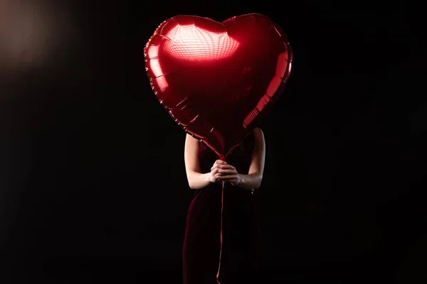 Frau im Kleid verschleiert Gesicht mit herzförmigem Luftballon im 14. Februar auf schwarzem Hintergrund — Stockfoto