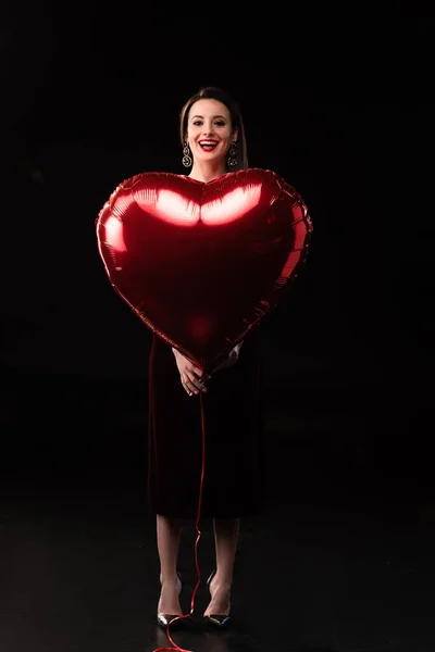 Mulher sorridente no vestido segurando balão em forma de coração no fundo preto — Fotografia de Stock