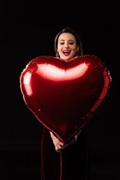 Shocked woman in dress holding heart-shaped balloon isolated on black — Stock Photo
