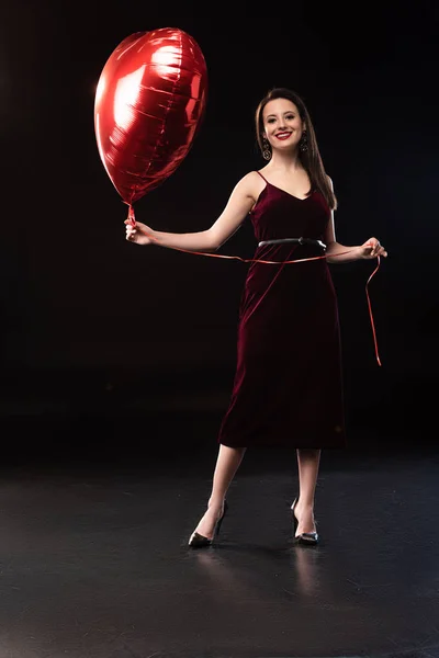 Mulher sorridente no vestido segurando balão em forma de coração no fundo preto — Fotografia de Stock