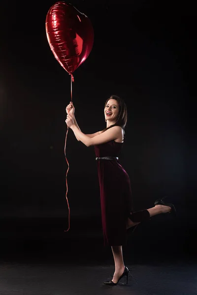 Vista lateral de mujer sonriente en vestido sosteniendo globo en forma de corazón sobre fondo negro - foto de stock