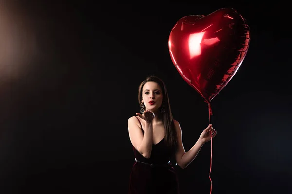 Женщина в платье держит воздушный шар в форме сердца и дует воздушный поцелуй на черном фоне — стоковое фото