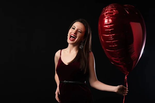 Mujer feliz en vestido sosteniendo globo en forma de corazón y gritando aislado en negro - foto de stock