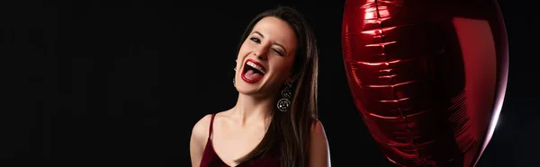 Panoramic shot of happy woman in dress holding heart-shaped balloon and screaming isolated on black — Stock Photo