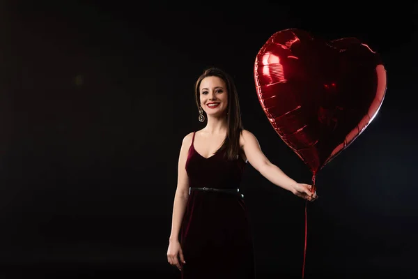 Mujer sonriente en vestido sosteniendo globo en forma de corazón aislado en negro - foto de stock