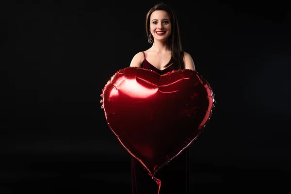 Усміхнена жінка тримає повітряну кулю у формі серця в 14 лютого ізольовано на чорному — стокове фото
