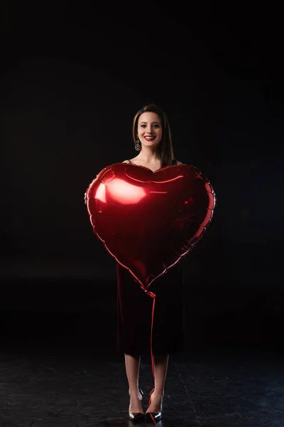 Sonriente mujer sosteniendo globo en forma de corazón en 14 febrero sobre fondo negro - foto de stock