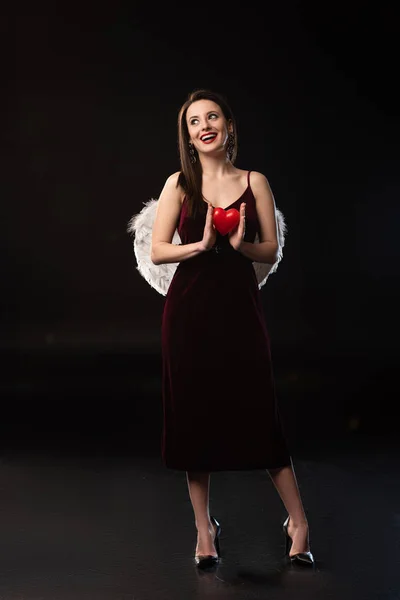 Mujer sonriente en vestido con alas sosteniendo modelo en forma de corazón en 14 febrero sobre fondo negro - foto de stock