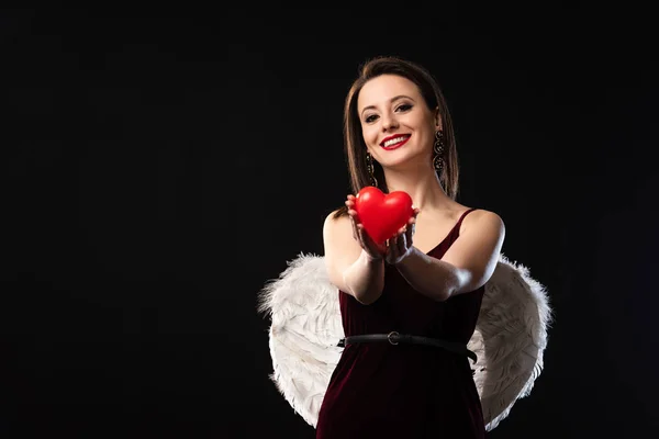 Mulher sorridente em vestido com asas segurando modelo em forma de coração em 14 de fevereiro isolado em preto — Fotografia de Stock